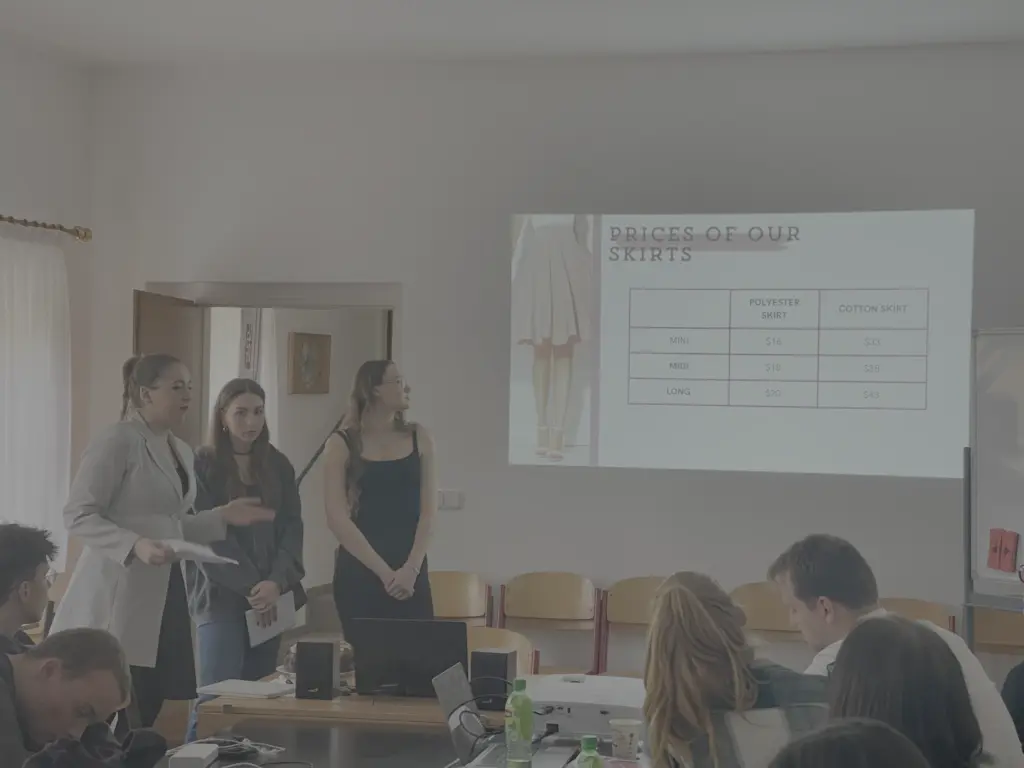 Tři žákyně prezentují svůj projekt spolužákům v učebně, na plátně anglický ceník výrobků